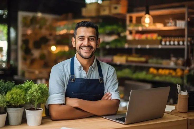 A smiling small business owner in an apron standing behind a counter with a laptop, in a shop lined with plants